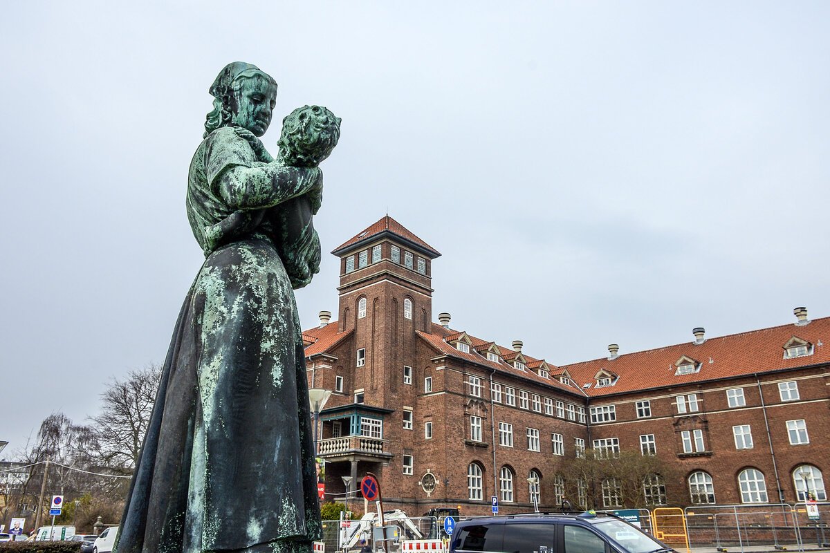 Vor dem Bispebjerg Hospital in Kopenhagen steht eine gr�n patinierte Bronzestatue, die eine Frau zeigt, die ein Kind liebevoll im Arm h�lt. Im Hintergrund erhebt sich das markante Backsteingeb�ude mit roten Ziegeld�chern und einem zentralen Turm, typisch f�r die Architektur des fr�hen 20. Jahrhunderts. Aufnahme: 24. M�rz 2026.