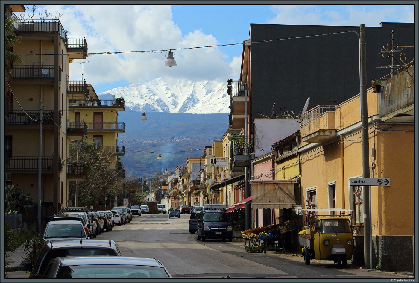 Blick entlang der Via Circumvallazione in der Kstenstadt Riposto auf den schneebedeckten tna. Rechts, an einem kleinen Lebensmittelgeschft steht einer der typischen lteren Ape-Transporter. (26.02.2024)
