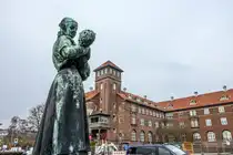 Vor dem Bispebjerg Hospital in Kopenhagen steht eine gr�n patinierte Bronzestatue, die eine Frau zeigt, die ein Kind liebevoll im Arm h�lt. Im Hintergrund erhebt sich das markante Backsteingeb�ude mit roten Ziegeld�chern und einem zentralen Turm, typisch f�r die Architektur des fr�hen 20. Jahrhunderts. Aufnahme: 24. M�rz 2026.