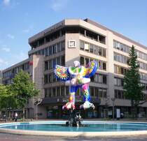 Auf der K� in Duisburg sieht man schon von weitem ,den 1991 - 1993 aus Polyester mit Teflonfarbbeschichtung errichteten 7,20 m hohen  Lifesaver -Brunnen von Niki de Saint Phalle. Der adler�hnliche Vogel, an den sich eine weibliche Figur klammert, steht mit ausgebreiteten Armen auf einem aus lackiertem Stahl aus Industrieabf�llen von Jean Tinguely errichteten Sockel, mit dessen Hilfe sich das Fabelwesen in der warmen Jahreszeit rhythmisch nach links und rechts bewegt und dabei Wasserkaskaden in den Brunnen spritzt.
