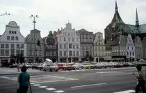 Rostock am 1. Juli 1990: Ernst-Th�lmann-Platz. - In den Jahren 1952 - 1991 hiess Neuer Markt, der historische Markt der Rostocker Mittelstadt, 'Ernst-Th�lmann-Platz'. Nach dem Zweiten Weltkrieg waren 90 Prozent der H�user hier zerst�rt, die Giebelh�user an der Westseite des Marktes - die H�user auf dem Foto - wurden aber neu errichtet. Rechts ist ein Teil der Marienkirche zu sehen.