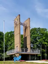 . Saarland - Das Europadenkmal in Berus in der Gemeinde �berherrn. 18.07.2014 (Jeanny)

Das Europadenkmal besteht aus zwei senkrecht aufragenden, 16 m hohen Betonplatten, die in ihrem unteren Teil eine Aussichtsplattform mit durchsichtigem Gel�nder durchbrechen. An ihren oberen Enden sind die beiden rechteckigen Pylone mittels eines Strahlenb�ndels aus mehreren Metallst�ben miteinander verbunden. Auf der Innenseite der beiden Pylone ist oberhalb der Plattform in Augenh�he eine Gedenktafel f�r die Politiker Konrad Adenauer, Robert Schuman und Alcide De Gasperi angebracht. Weitere Plaketten f�r Helmut Bulle, Josef Besch und Paul Henri Spaak wurden nach der Einweihung angebracht.

Das Europadenkmal ist heute das Wahrzeichen der Grenzgemeinde �berherrn. Seine Lage und die Aussicht machen das Denkmal zum beliebten Haltepunkt auf Spazierg�ngen und Ausfl�gen. Die Stirnseite des Denkmals ist ungef�hr in Richtung auf Saarbr�cken orientiert, was auf die uralte Br�ckenfunktion des Saarraumes als Kreuzungspunkt von Fernwegen und auf seine weit j�ngere Br�ckenfunktion im deutsch-franz�sischen Verh�ltnis verweist. Gerald Motsch versuchte, die deutsch-franz�sische Geschichte im Laufe der Jahrhunderte in vertikaler Form durch die beiden Platten ins Bild zu setzen. Die horizontale Plattform des Denkmals signalisiert die gemeinsame historische Wurzel der beiden Nationalstaaten im keltisch, romanisch und germanisch gepr�gten fr�hgeschichtlichen Kulturraum. W�hrend die Betonplatten au�er im Untergrund und in der Plattform nirgends verbunden sind, wurde in ihrem oberen Teil durch die geb�ndelten St�be eine Verbindung aus Metall, d.h. aus einem anderen Material geschaffen.

Der blaue Hirsch im Vordergrund erinnert an das Jahr 2007, als Luxemburg gemeinsam mit der Gro�region europ�ische Kulturhauptstadt war. Er diente als Logo dieses Ereignisses. 